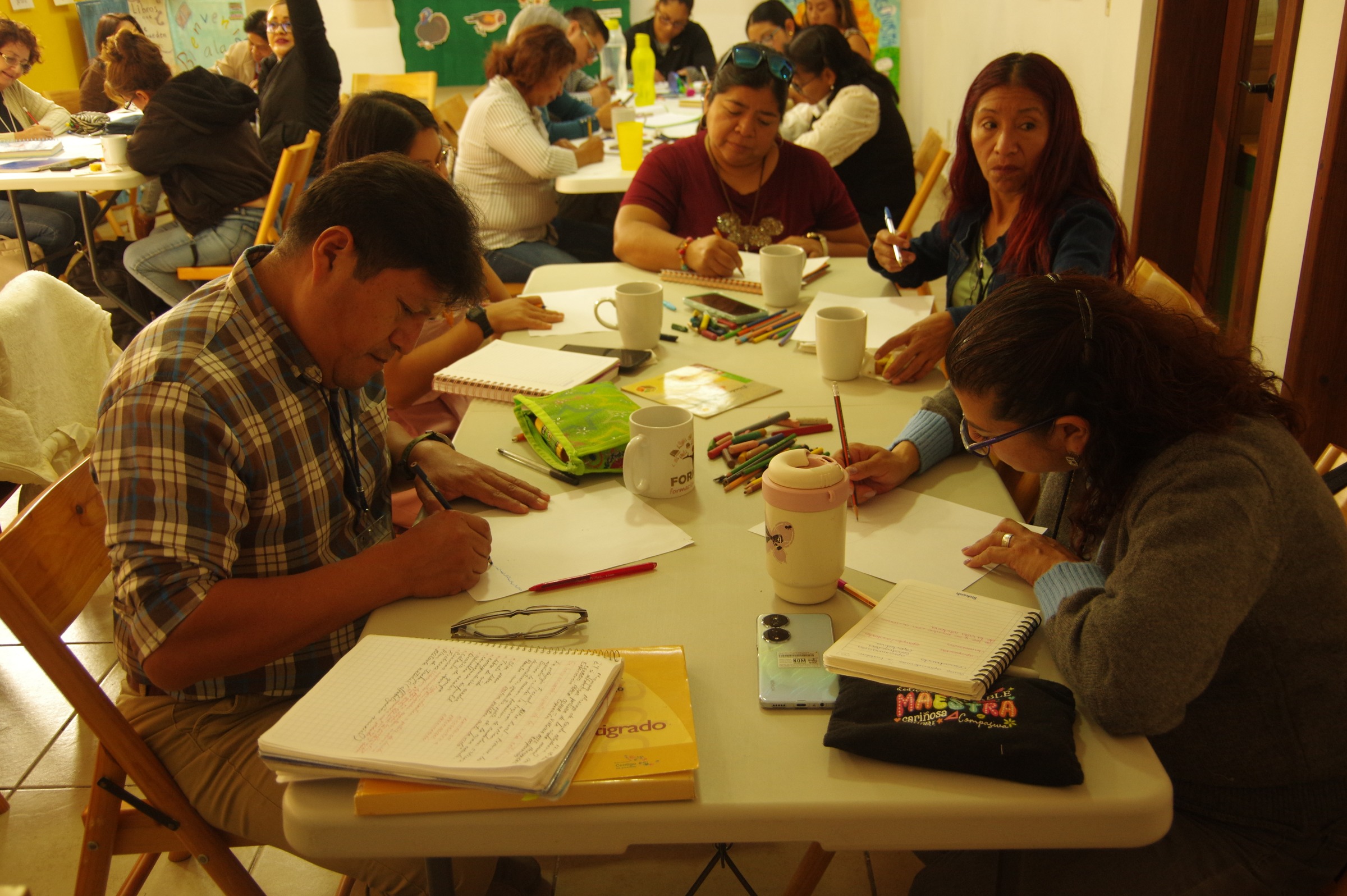 Personas escriben y revisan cuadernos alrededor de mesas.